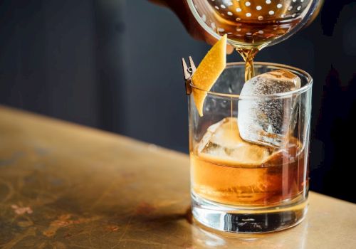 A rocks glass with whiskey and ice being topped with a craft pour, garnished with an orange twist and a small wooden pick.