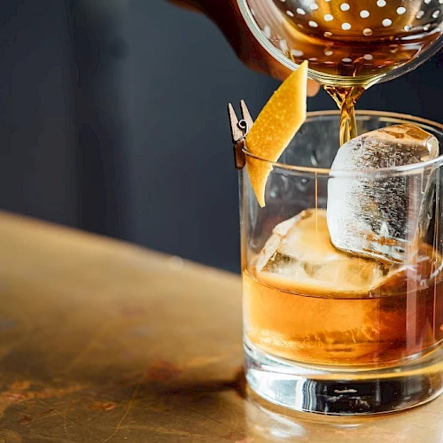 A rocks glass with whiskey and ice being topped with a craft pour, garnished with an orange twist and a small wooden pick.