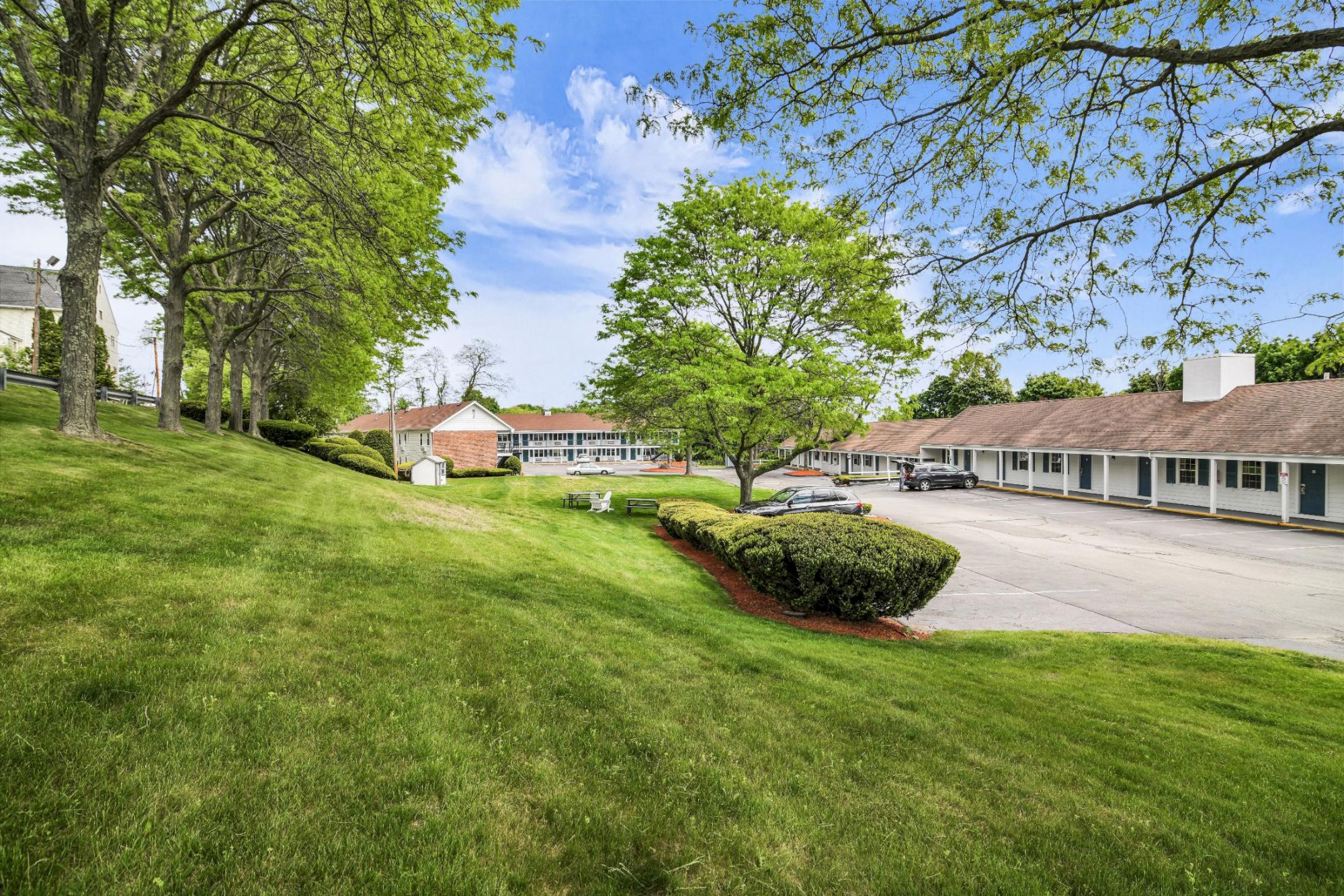 A serene suburban motel or campus area with single-story buildings, green lawns, trees, and a curved driveway under a bright blue sky.