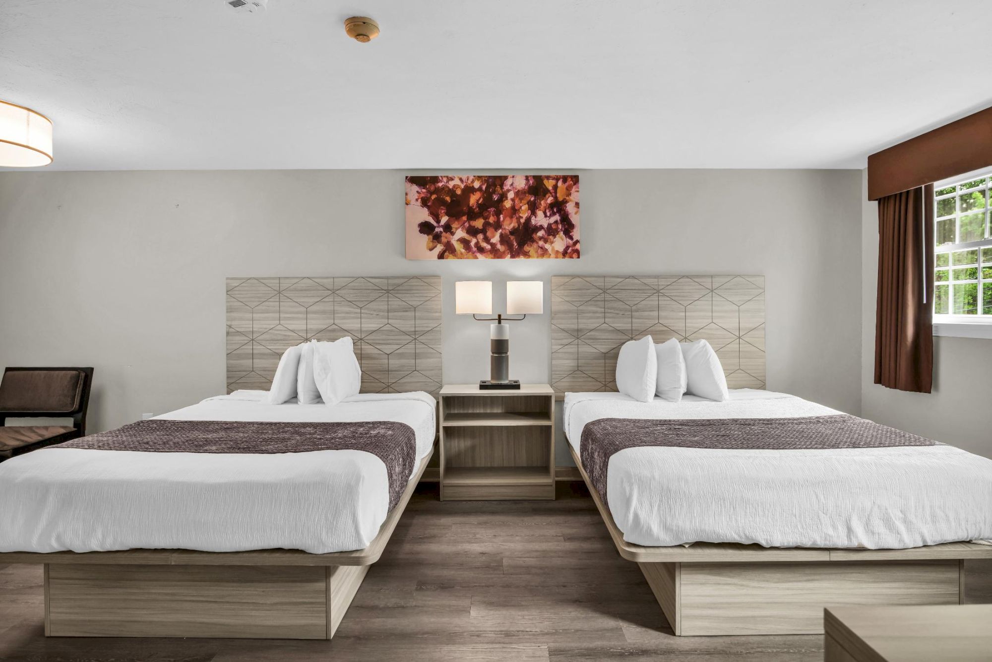 Two neatly made hotel beds with matching wooden headboards, a shared nightstand, lamps, and a framed art piece, in a bright, minimalist room.