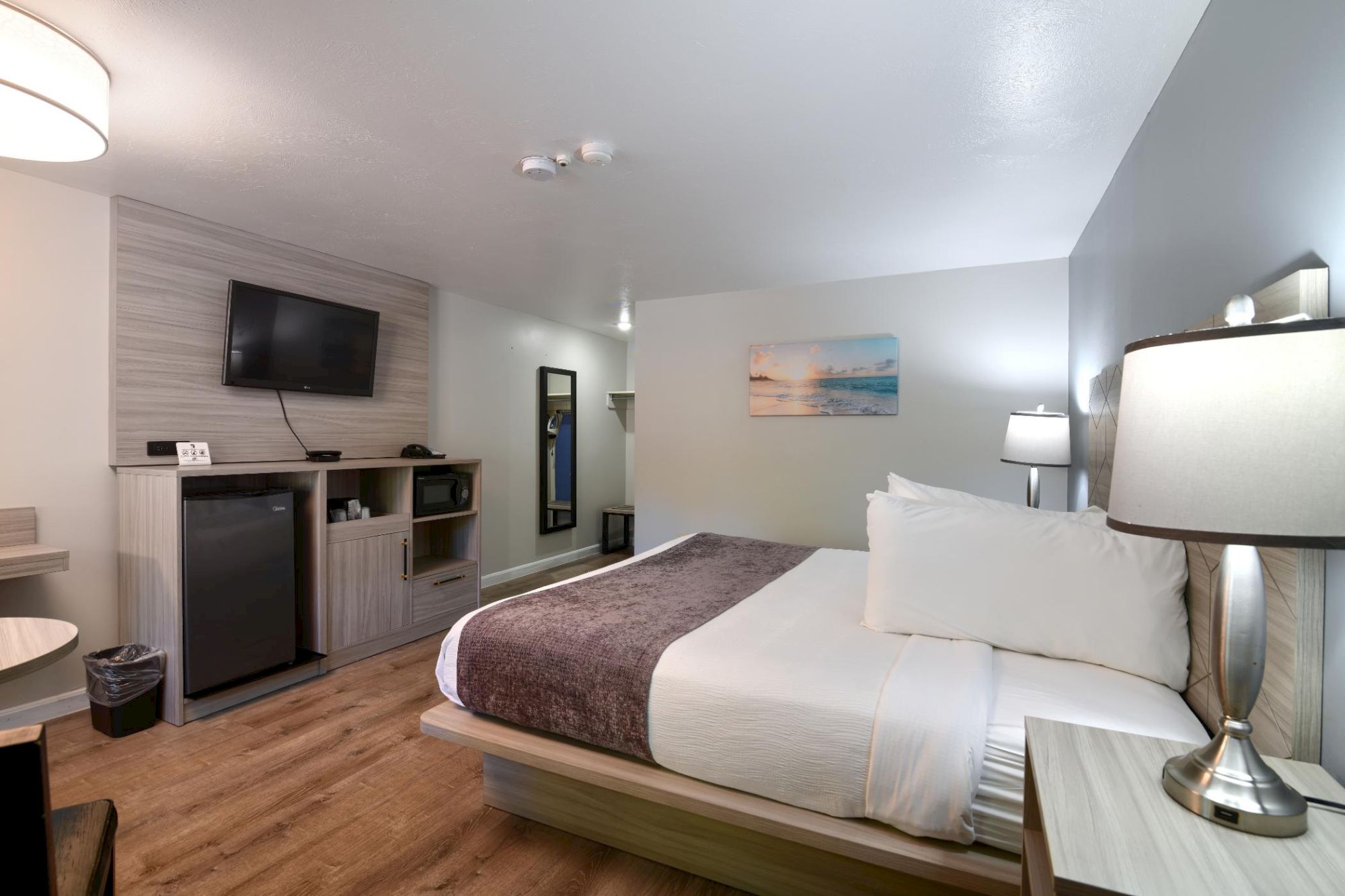 A modern hotel room with a king bed, bedside lamps, a small desk, a TV on a wood-paneled wall, a minibar, and soft blue-gray decor.