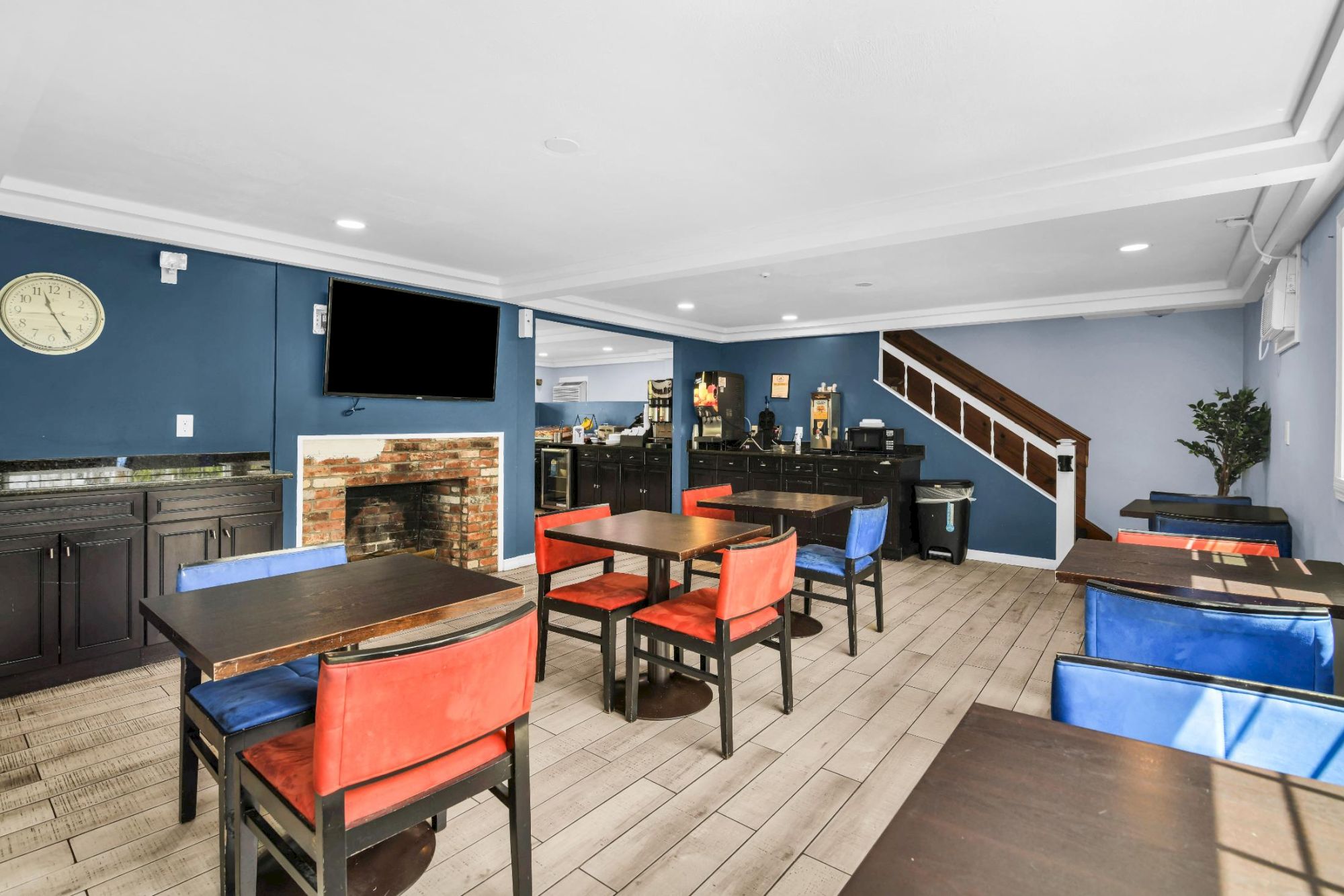 A cozy cafe or lounge with blue walls, red and blue chairs, wooden tables, a TV, a fireplace, and a staircase in the back, plus a potted plant near the stairs.