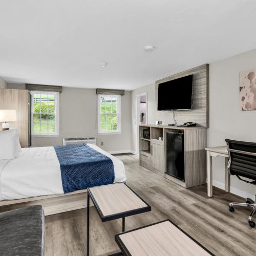 A modern hotel room with two beds, a small seating area, desk with lamp, wall-mounted TV, and a mini kitchenette. The decor is light and contemporary, with neutral tones and wood accents.