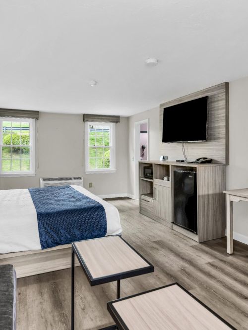 A modern hotel room with two beds, a small seating area, desk with lamp, wall-mounted TV, and a mini kitchenette. The decor is light and contemporary, with neutral tones and wood accents.