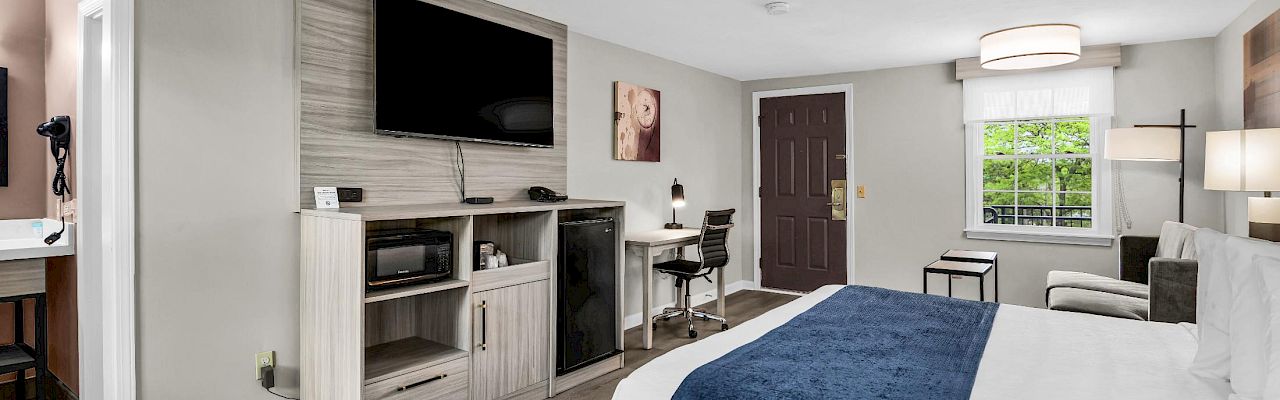A modern hotel room with a king bed, blue throw, a wall-mounted TV, a desk, a small sofa, and a kitchenette area in soft gray tones.