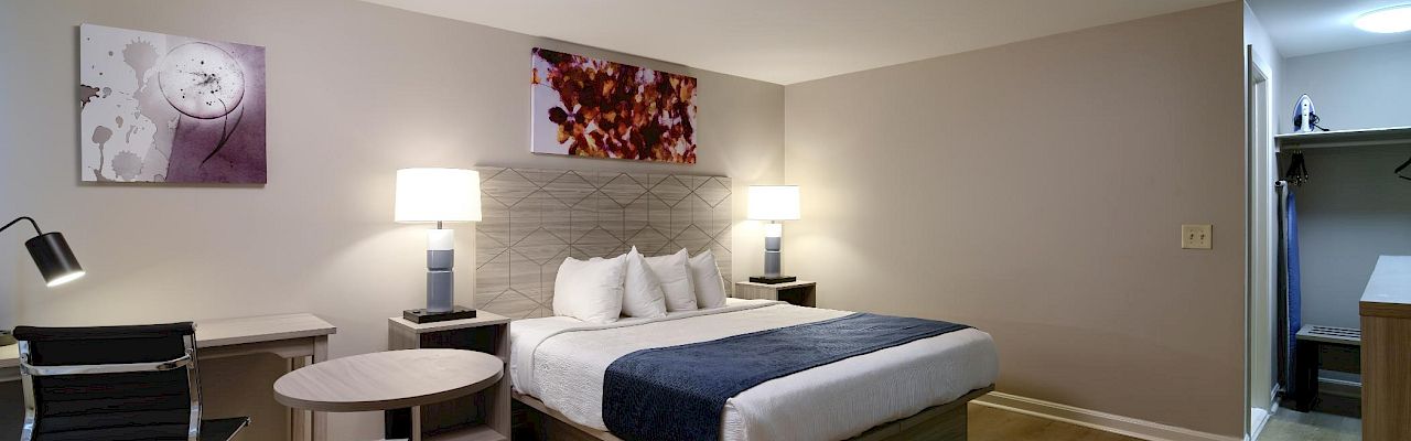 A modern hotel room with a king bed, two nightstands with lamps, a seating area, and wall art; doorway to a small closet area on the right.