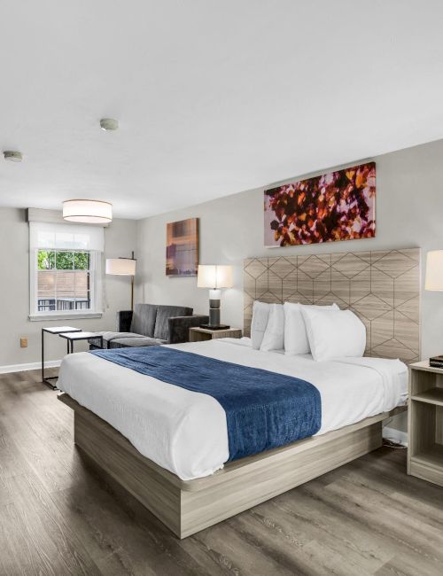 A modern hotel room with two queen beds, neutral tones, wooden accents, a work desk, flat screen above a bench, and an open closet area with hangers, all in a bright, spacious layout.