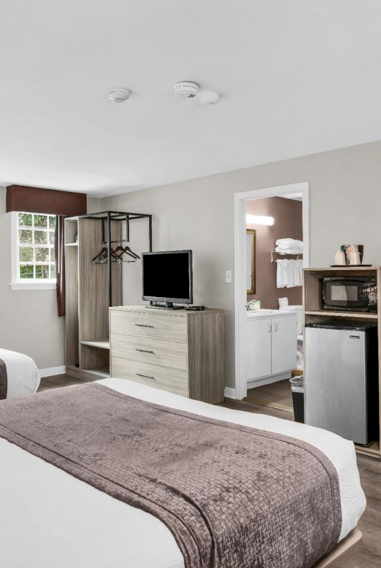A clean hotel room with two beds, a desk, TV, mini-fridge, and a small seating area; neutral decor and practical furnishings.