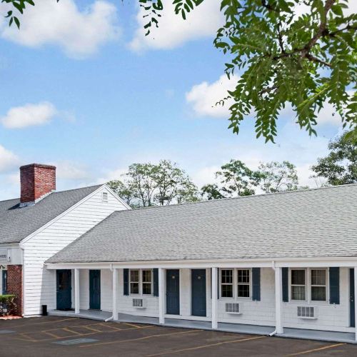 A single-story storefront with a long white fa&ccedil;ade, a gray roof, and a row of windows, set beneath a bright blue sky with green trees.