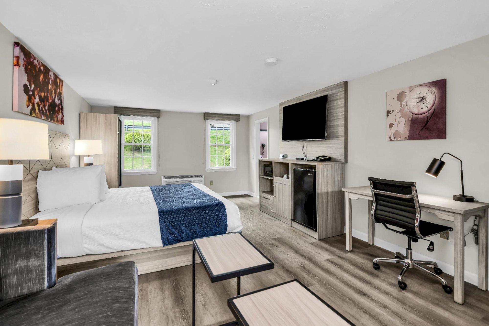 A modern hotel room with two beds, a desk and chair, a wall-mounted TV above a mini-fridge, and a small seating area by a pair of windows.