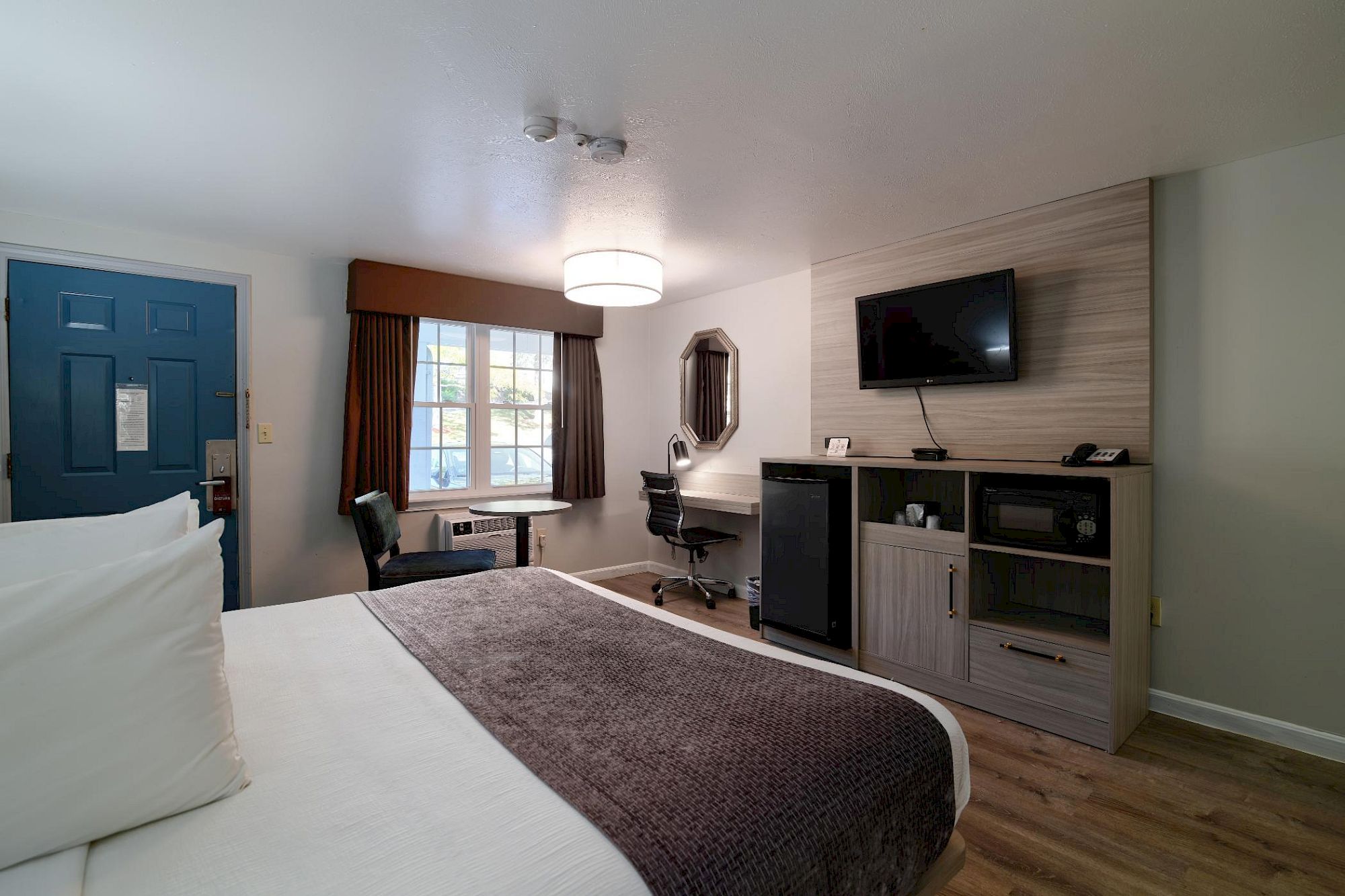 Hotel-style room with a bed, desk, seating area, and a mounted TV; wood floors, blue door, and a modern mini-fridge cabinet.