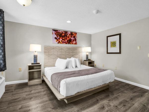 A modern hotel room with a king bed, two nightstands, lamps, a framed wall art piece, and a neutral color scheme with wood flooring. Top it at 140 characters, always ending the sentence.
