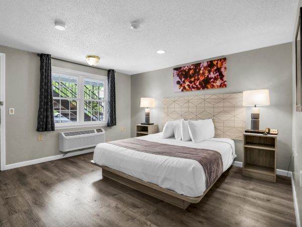 A modern hotel room with a large bed, two nightstands, lamps, a window with curtains, wood flooring, a wall art piece, and a serene, neutral color palette.