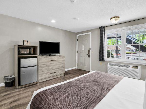 A simple hotel-style hotel room: bed in foreground, dresser with TV, mini-fridge, desk area, window with curtains, and an AC/heat unit by the window.
