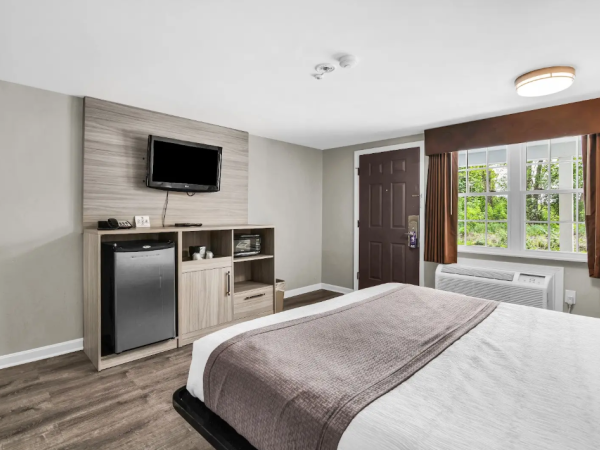 A tidy hotel-style bedroom with a queen bed, small TV cabinet, and a window with curtains and an air conditioner.
