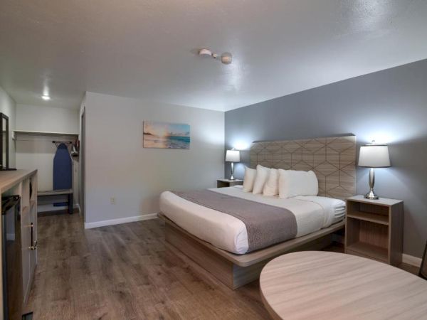 A modern hotel room with a king bed, two nightstands, soft lighting, a round table, a small desk, and an open closet area in the corner.