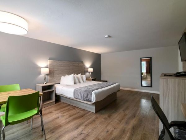 A modern hotel room with a large bed, two nightstands, a wall-mounted TV, wooden floor, a small desk with green chairs, and a mirror across from the bed.