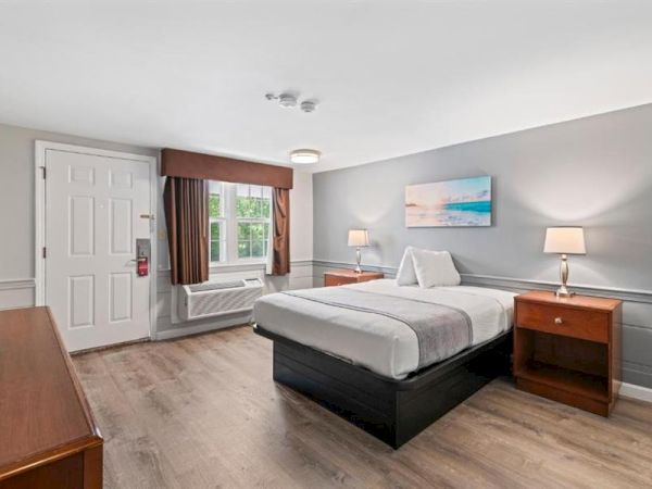 A bright hotel-style room with a king bed, two wooden nightstands, lamps, a dresser, window with curtains, and light gray walls.