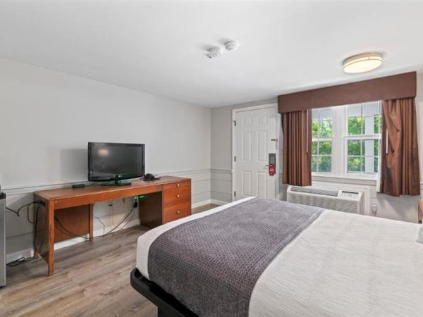 A hotel-style bedroom with two beds, a wooden desk and TV, a small fridge, window with brown curtains, and an air conditioner, clean and simple.