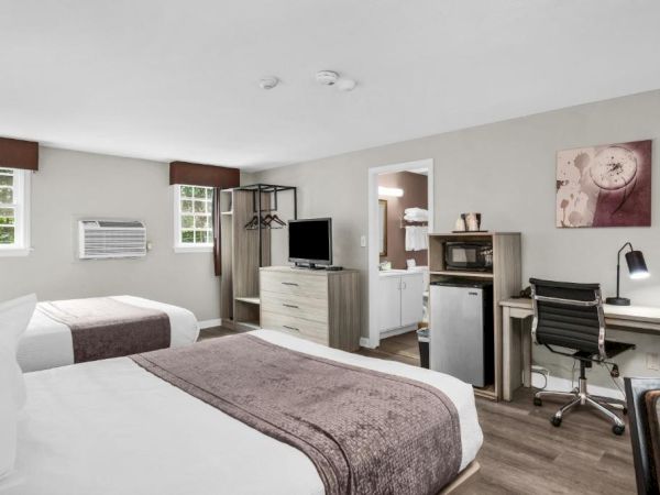 A bright hotel room with two neatly made beds, a desk and chair, a small TV, dresser, mini-fridge, microwave, and a seating area near a doorway.