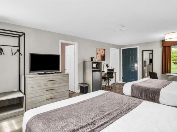 A bright hotel room with two beds, a dresser, TV, open closet, desk, and a window with brown curtains. The space looks neat and modern.