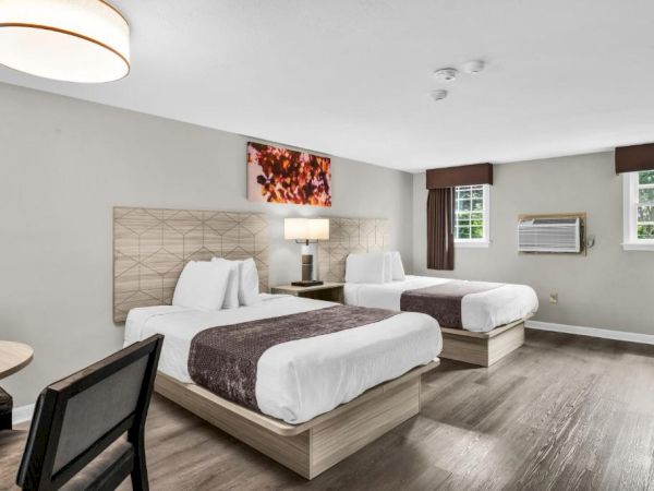 Modern hotel room with two queen beds, neutral decor, a wall TV, desk chair, and large windows with brown curtains.