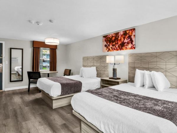 A modern hotel room with two queen beds, neutral decor, a full-length mirror, a door to the hallway, a small desk, and a lit bedside lamp, with a red wall art piece.