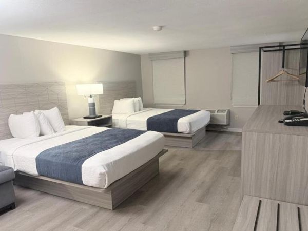 A clean, modern hotel room with two queen beds, a desk area, a wall-mounted TV, nightstand lamps, and a dresser; minimalist and bright.