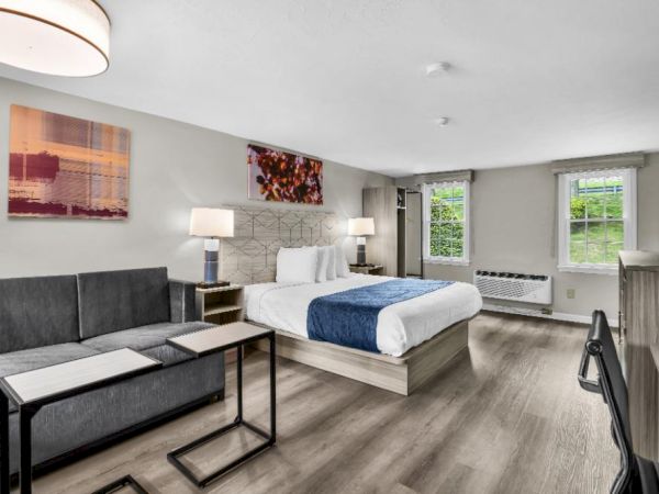 A modern hotel suite with a king bed, grey sofa, side tables, lamps, and artwork; kitchenette area and large windows brighten the wooden floor.