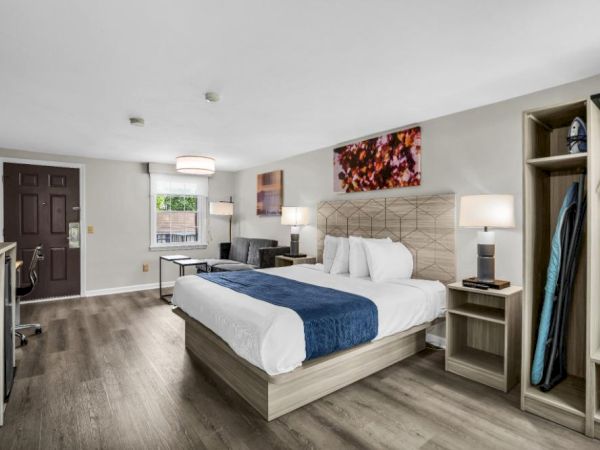 A modern hotel room with a king bed, neutral tones, wood floors, a desk, seating area, open closet, and contemporary art above the headboard.