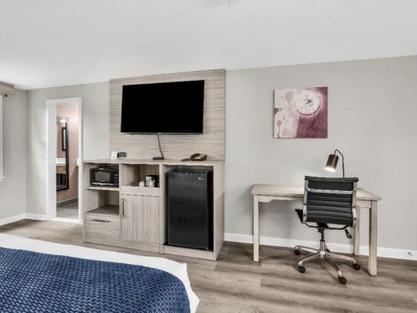 A modern hotel/guest room with a TV on a stone accent wall above a cabinet, a mini fridge, a desk with a chair, and a blue bedspread in the foreground.