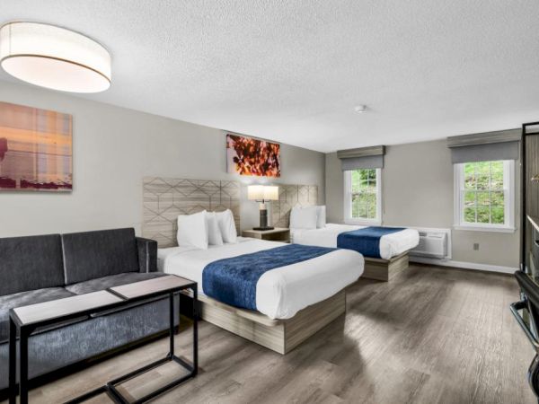 A bright hotel room with two queen beds, a gray sofa, a small desk, wall art, and a flat-screen TV; neutral tones, wood flooring, and natural light.