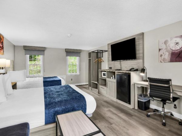 A bright hotel room with two beds, a desk, TV, and a small kitchenette area, in a modern, minimalist style.