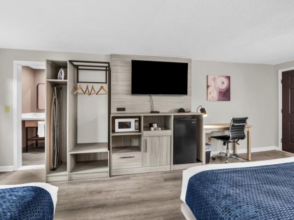 A hotel-style hotel room with two beds, an open wardrobe, a TV on a console, a small fridge, a desk and chair, and a doorway to a bathroom, all in neutral tones.