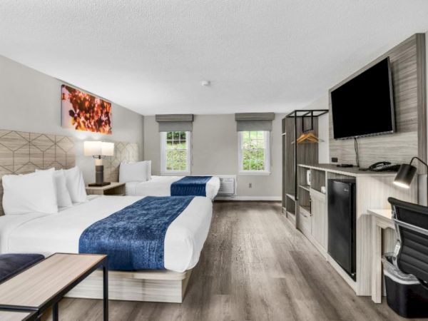 A modern hotel room with two queen beds, a desk, sofa, flat-screen TV, mini-fridge, and an open wardrobe, all in a bright, airy space.