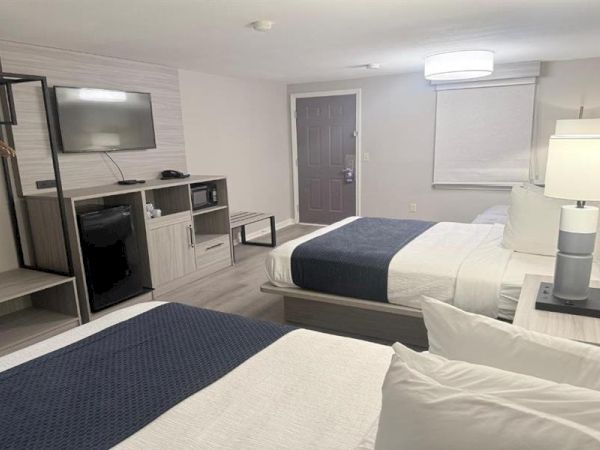 Two neatly arranged hotel rooms with two beds, a desk, TV, mini-fridge, wardrobe, and modern lighting in a clean, gray-blue themed space.