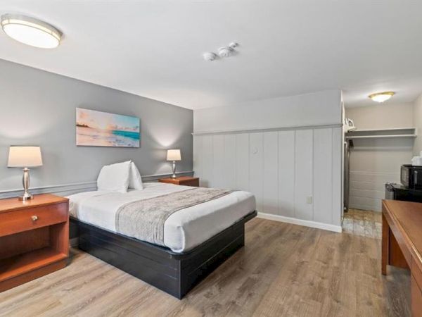 A motel-style hotel room with a large bed, two bedside tables, lamps, a dresser, a small open closet, and a desk area with a coffee maker in a bright, clean space.
