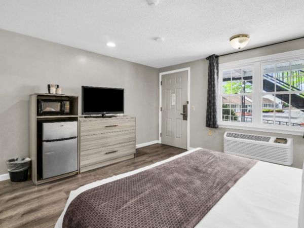 A tidy hotel-style room with a bed, a dresser, a mini-fridge, a TV, a windowed wall unit, and neutral decor, a door, and soft lighting.