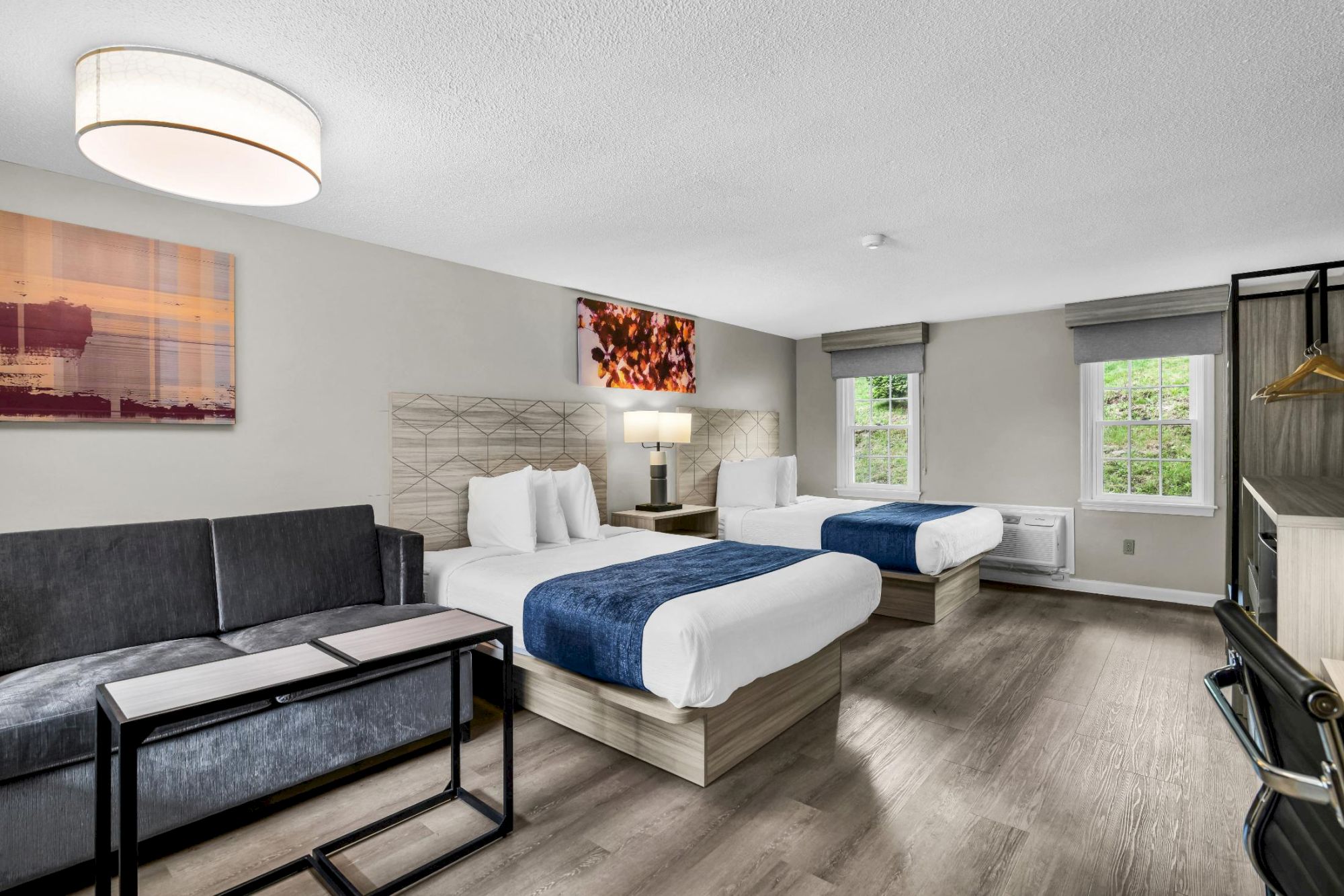 A modern hotel-style bedroom with two beds, lamps, blinds, and large windows, styled in neutral tones and blue accents.