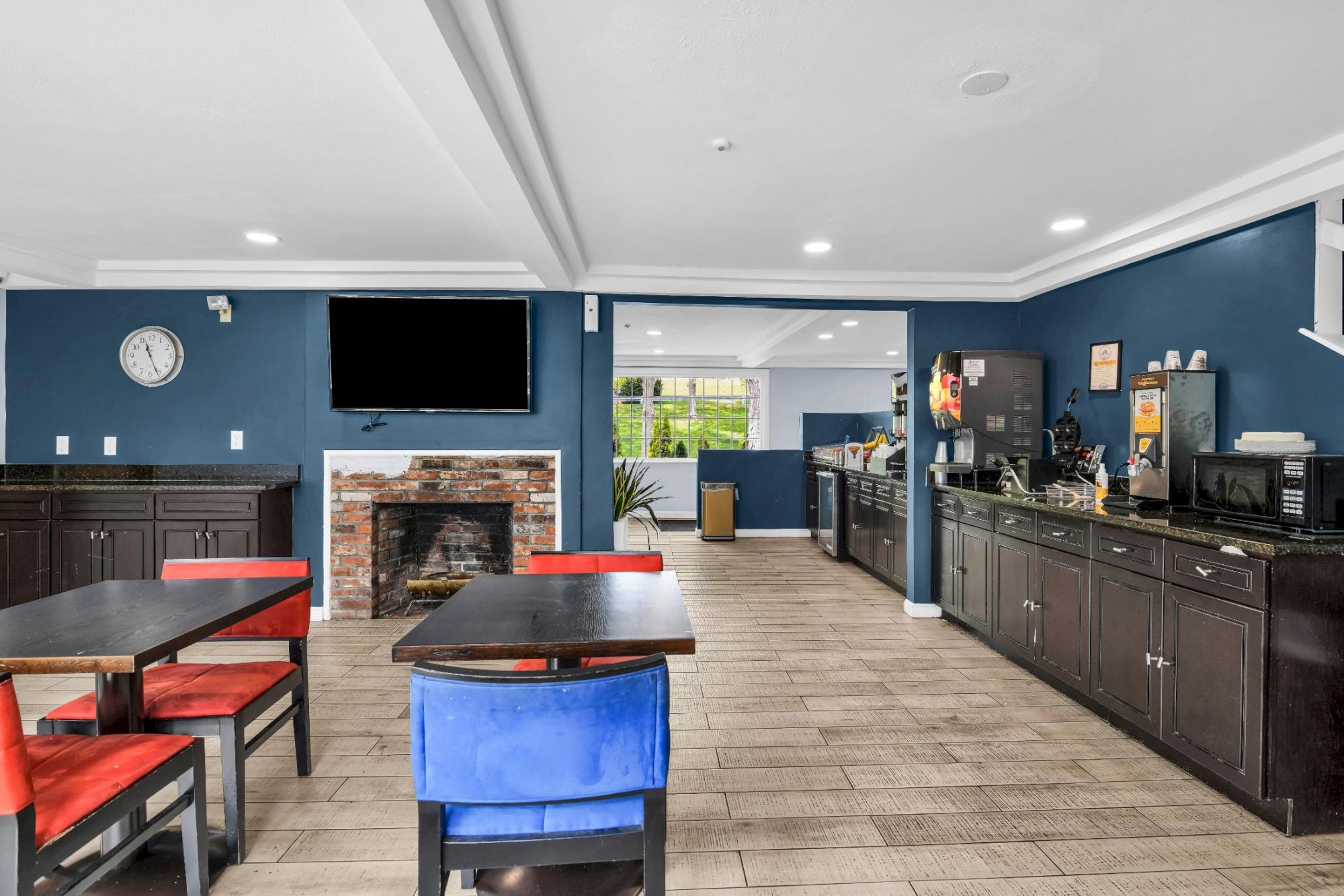 A cozy communal game room with a blue accent wall, a fireplace, a mounted TV, red-and-black chairs, and a long counter with coffee machines and cabinets along the right wall.