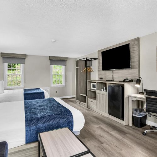 A modern hotel room with two beds, a desk and chair, a flat-screen TV, a small fireplace, kitchenette area, and a minimalist gray-blue decor with natural light.