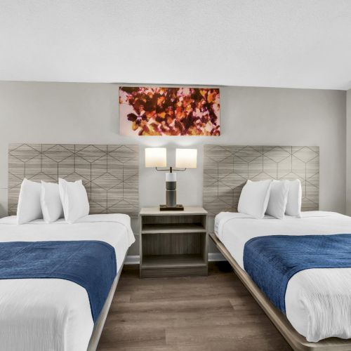 Two double beds with white linens and navy blue throws sit side by side in a bright hotel room, sharing a nightstand and framed art above.