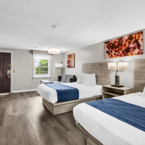 A modern hotel room with two queen beds, a desk, flat-screen, neutral walls, and warm wood floors, plus artwork and a doorway to the hall.