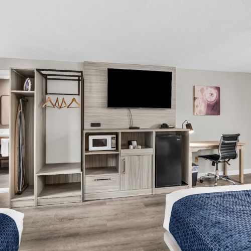 A hotel-style room with two beds, a TV, open closet, a small kitchenette, a desk and chair, a mini-fridge, and neutral decor with a doorway on the left.