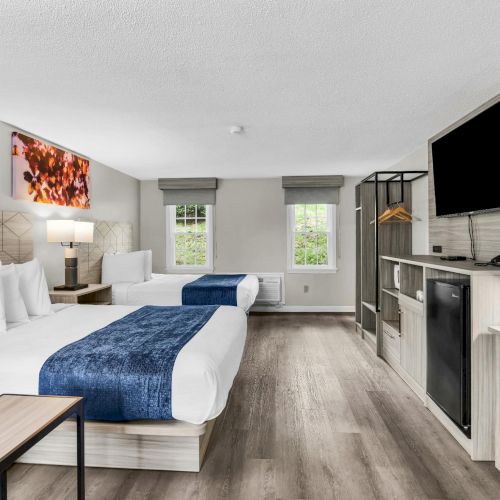 A modern hotel room with two queen beds, a desk, flat-screen TV, mini fridge, a sofa, and a coffee table, bright windows, and gray wood flooring.