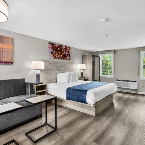 A modern hotel suite with a king bed, gray sofa, and a small work desk; warm lighting, abstract art, and large windows in a bright, airy room.