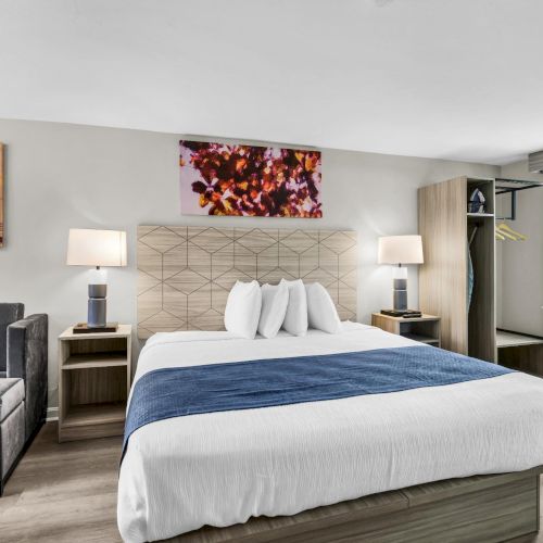 A modern hotel room with a king bed, blue throw, two bedside tables with lamps, a gray sofa, artwork above, and an open closet by a window.