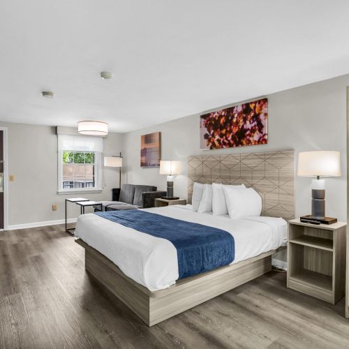 A modern hotel room with two queen beds, neutral decor, wood flooring, nightstands, a desk, a seating area, open closet, and artwork above the bed.