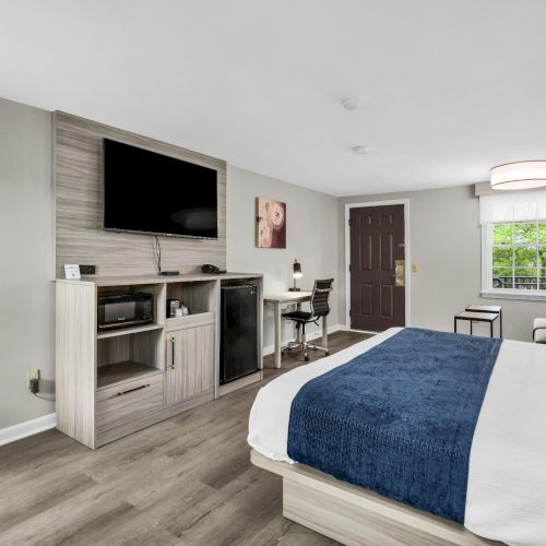 A modern hotel room with a bed, blue throw, seating area, desk, and a flat-screen TV on a console against a light gray wall.