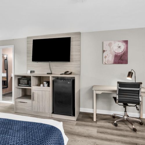 A modern hotel-like bedroom with a wall-mounted TV, a small kitchenette with a mini-fridge, a desk and chair, and a blue bedspread, bright and airy.
